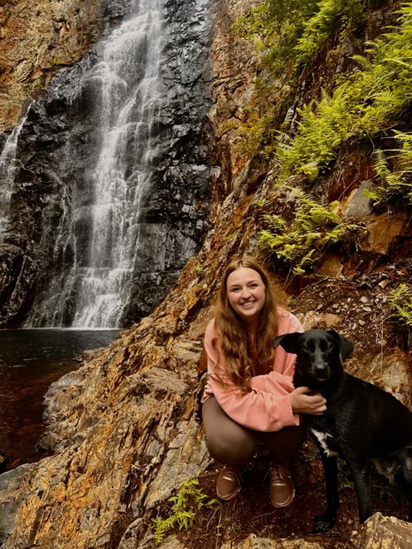 Woman crouching on rocky terrain beside a cascading waterfall with a black dog by her side, outdoors.